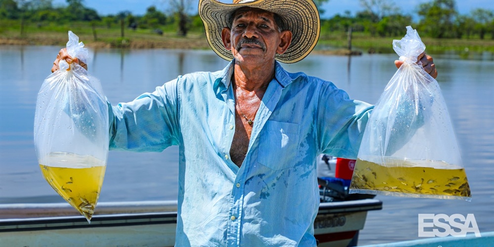 Hombre con sombrero en su cabeza y sosteniendo dos bolsas de peces, de fondo el río
